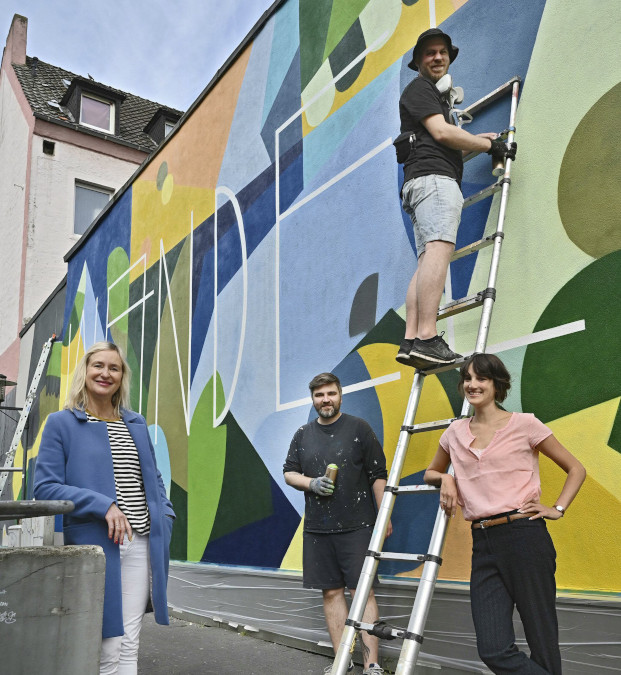   Künstlerduo Clemens Brück und Sebastian Saffenreuter (mit Hut) zusammen mit Claire Neidhardt und Christiane Gabbert am Stadtmarkt in der Innenstadt vor einer bunten Graffitiwand.